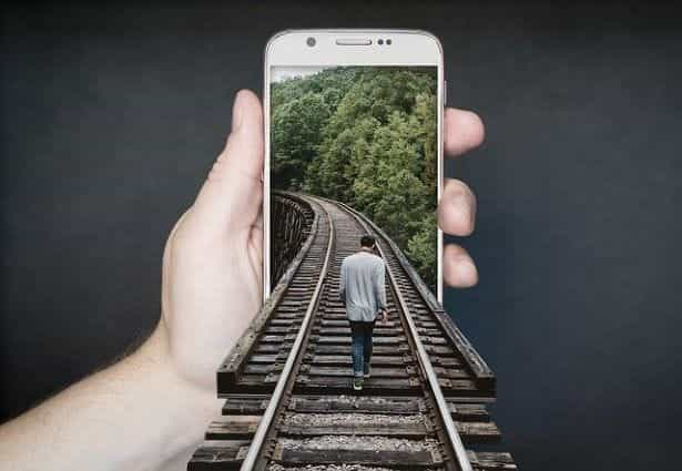 A person holding up a smartphone which has a man walking along train tracks emerging out of it.