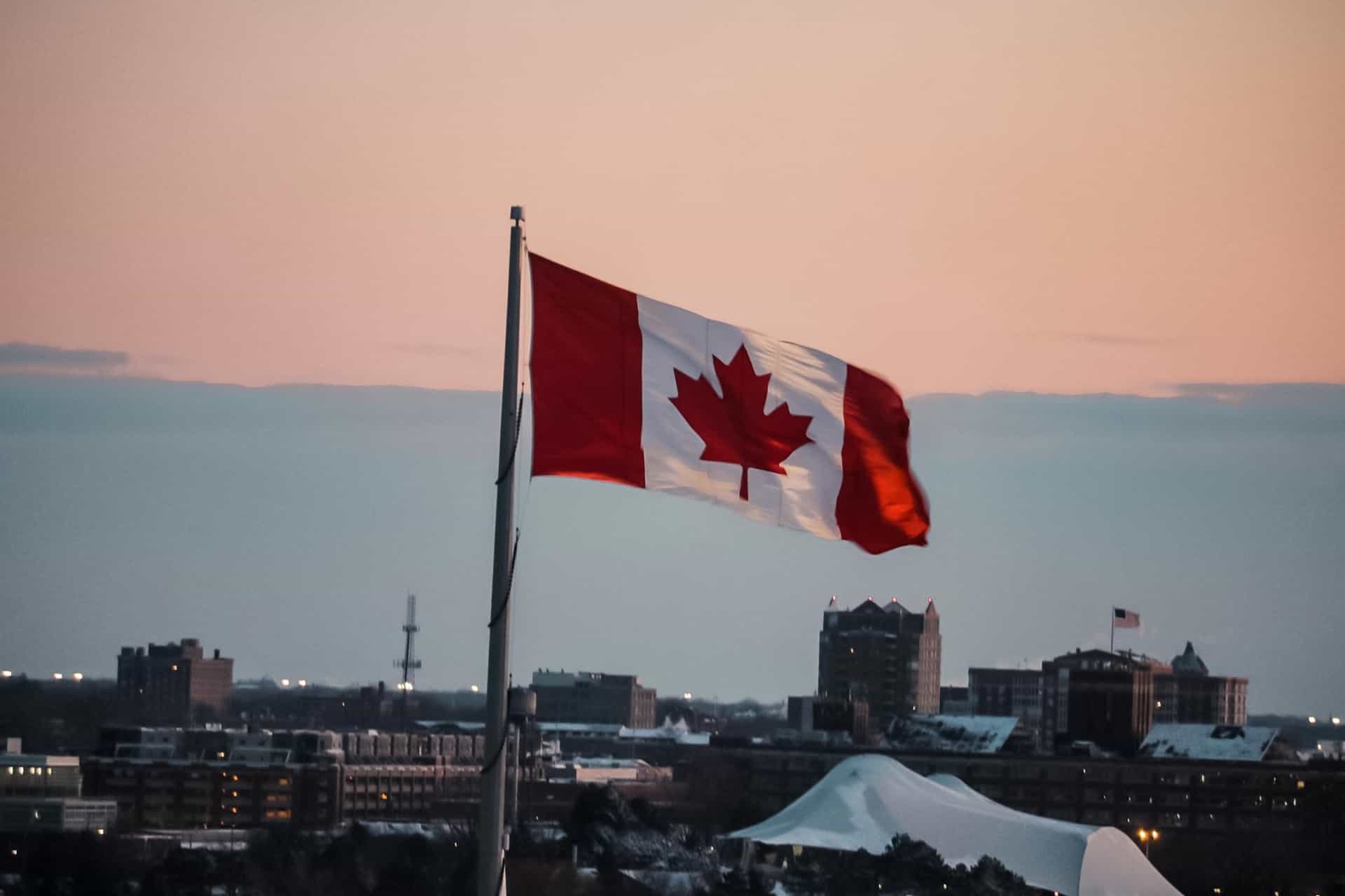 The Canadian national flag, featuring its iconic red and white colors and a Maple Leaf, flying high above a city in the country.